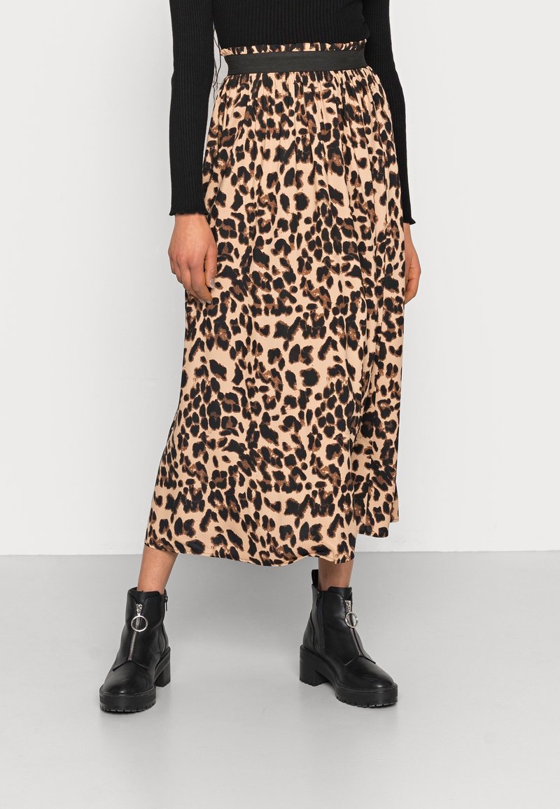 Femme portant une jupe midi à imprimé léopard avec une ceinture noire, un haut à manches longues côtelé noir, et des bottines noires avec des anneaux de fermeture éclair argentés.