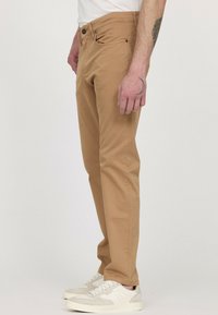 Chinos kaki, coupe slim, devant plat avec passants de ceinture, cinq poches et marquage subtil. Portés avec des baskets blanches, en tissu de coton doux.