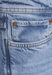 Ljusblå denimtyg med strukturerad yta. Har en bakficka med bruna nitar och orange sömmar längs sömmarna.