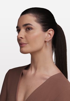 Žena s tamnom kosom u repu nosi smeđu majicu sa V-izrezom i ružičasto-zlatnu geometrijsku naušnicu s malim dragim kamenom.