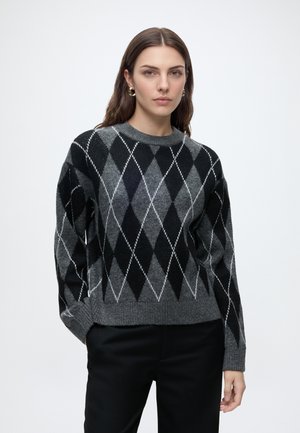 Femme aux longs cheveux bruns portant des boucles d'oreilles créoles dorées et un pull gris et noir à motif losanges avec un pantalon noir sur fond blanc.