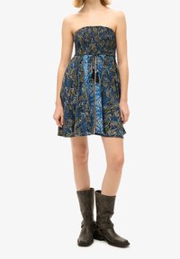 Robe bleue sans bretelles à motif cachemire avec un haut smocké et un panneau floral central, portée avec des bottes en cuir marron foncé mi-mollet avec des clous et des sangles.