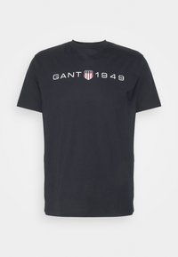 Camiseta de algodón negra con cuello redondo, mangas cortas y un gráfico blanco "GANT 1949" con un emblema de escudo en rojo y blanco.