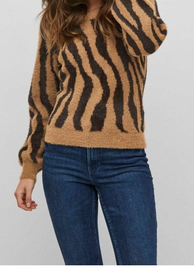 Femme portant un pull marron en laine à motifs d'animaux noirs et un jean en denim bleu, debout devant un fond clair uni.