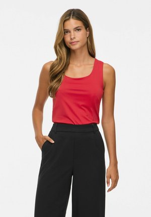 Femme aux longs cheveux portant un haut rouge sans manches et un pantalon noir taille haute, debout avec une main dans la poche.