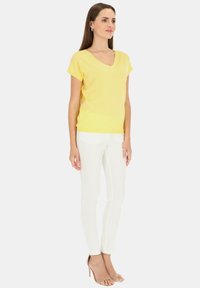 Żółta bluzka z krótkim rękawem i dekoltem w kształcie litery V, wykonana z lekkiego materiału, w połączeniu z białymi obcisłymi spodniami i cielistymi sandałami na wysokim obcasie.
