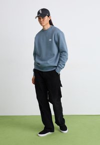 Felpa azzurra chiara con polsini a costine, abbinata a pantaloni di velluto a coste neri e scarpe slip-on nere. Cappellino nero con logo, in piedi su un pavimento verde.