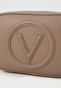 Ruda odinė rankinė su tekstūruotu paviršiumi, iškilniu apvaliu logotipu su iškiltu "V" ir matomomis siūlėmis aplink kraštus.