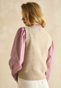 Beige Strickweste über einer pink-weiß gestreiften Bluse mit Puffärmeln. Weicher Stoff, runde Halslinie und gerippter Saum.