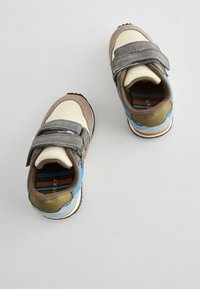 Lichte atletische schoenen met een mix van beige, grijs, blauw en groene materialen, voorzien van een klittenbandsluiting en gestructureerde oppervlakken.