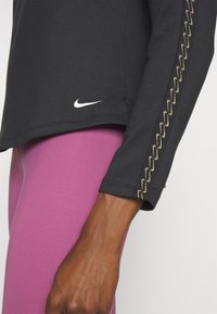 Zwarte langemouwen top met gouden gebogen stiksel op de mouw en wit Nike-logo. Draagt met roze leggings. Glad textiel.