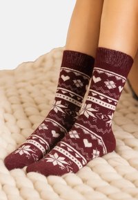 Legs wearing maroon woolen socks with white heart and snowflake patterns, resting on a chunky cream knit blanket.