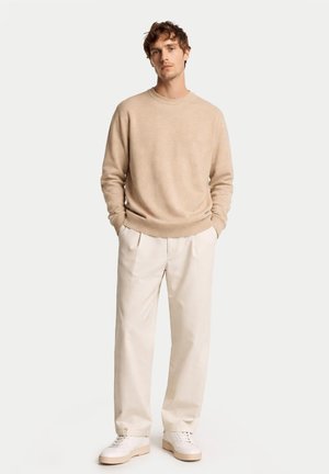 Homme portant un pull beige, un pantalon ample blanc cassé et des baskets blanches, debout les mains dans les poches devant un fond uni.