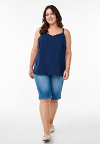 Top sin mangas azul marino con escote en forma de concha, combinado con pantalones cortos de mezclilla bermuda de un azul claro. La modelo lleva zapatos planos beige. Tela suave, diseño casual.