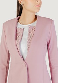 Donna che indossa un blazer rosa chiaro sopra una maglietta bianca con decorazioni in perle rosa e bead d'argento vicino al colletto.