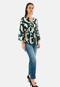 Blusa holgada con un audaz patrón geométrico en colores verde, negro y blanco. Combinada con jeans ajustados azules y tacones negros.