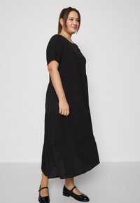 Crna, kratkih rukava, višeslojna maksi haljina s okruglim izrezom. Glatka tkanina s lepršavim siluetom. Crne lakirane kožne cipele upotpunjuju look.