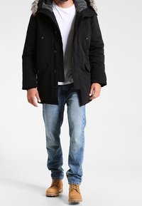 Parka noire avec capuche bordée de fausse fourrure, fermetures à boutons et deux poches latérales, portée sur une chemise blanche et un jean bleu, accompagnée de bottes couleur tan.