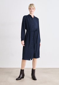 Navy blue button-up dress with long sleeves, tie waist, and side slit. Smooth fabric texture, paired with brown ankle boots.