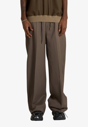 Persona con pantaloni marroni larghi con coulisse e scarpe da ginnastica nere, in piedi con le mani rilassate lungo i fianchi su sfondo bianco.