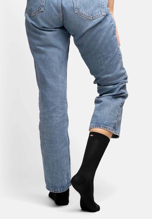Chaussette noire avec un logo rouge et blanc, portée avec un jean en denim bleu, présentant une coupe décontractée et des poignets légèrement retroussés.