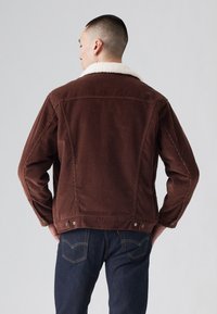 Bruine corduroy jas met een witte faux schapenvacht kraag. Kenmerken zijn knoopsluitingen en een getailleerde pasvorm. Spijkerbroeken zichtbaar bij de taille.