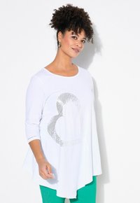 Weißes Longsleeve mit Rundhalsausschnitt und einem silbernen Herzdesign. Der Stoff scheint leicht zu sein, mit einer fließenden Form und abgerundetem Saum.