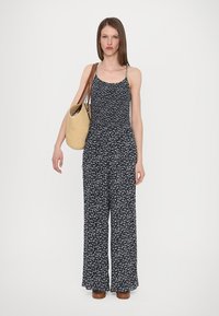 Giubbotto floreale di colore blu navy con spalline sottili e pantaloni larghi; il modello tiene una borsa di paglia con spalline marroni.