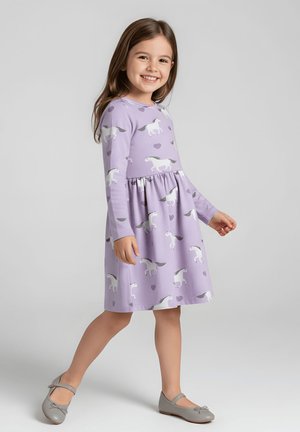 Młoda dziewczyna uśmiecha się, nosi lawendową sukienkę z długim rękawem z białymi wzorami koni i serc oraz szare buty Mary Jane, spaceruje po szarym tle.