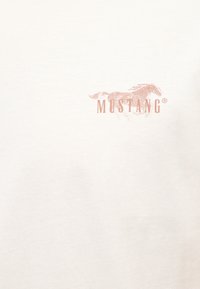 Logo Mustang avec cheval au galop et texte en majuscules, imprimé en marron clair sur tissu écru.