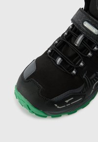 Sapatilha atlética preta e verde com atacadores elásticos, uma strap de Velcro e detalhes refletivos na ponta e nos lados.