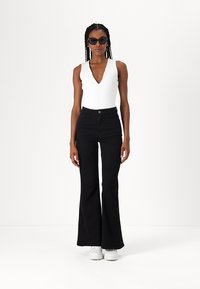White sleeveless bodysuit paired with high-waisted black flared jeans. Model wears dark sunglasses and white sneakers. Minimalist design.