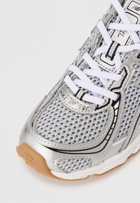 Baskets de sport argentées avec un dessus en maille, lacets blancs, semelle en caoutchouc et un logo proéminent dans un design texturé. Accentué par des lignes noires.