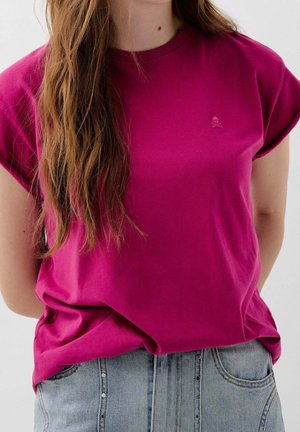 Woman with long brown hair wearing a magenta T-shirt with a small skull embroidery and light blue denim jeans, standing against a plain background.