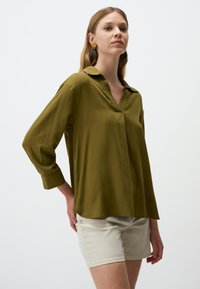Blusa leggera a maniche lunghe di colore verde oliva con scollo a V e colletto pieghettato. Abbinata a pantaloni corti color crema, indossata con accessori neutri.