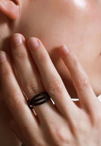 Zwarte ring met twee overlappende ronde banden om een hand, met natuurlijke, onafgebeten nagels en lichtjes gestructureerde huid tegen een neutrale achtergrond.
