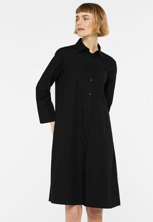 Frau mit kurzen blonden Haaren, die ein schwarzes, knielanges Kleid mit langen Ärmeln, Kragen und Knopfleiste trägt, schaut nach links, die Hände hinter dem Rücken.