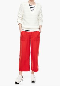 Pull blanc en maille avec col en V, associé à un pantalon rouge à jambes larges avec de grandes poches, et des baskets blanches avec des accents foncés.