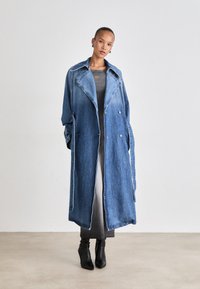 Denim trenchcoat in een lichte naar donkere blauwe gradient, met een dubbelzijdig ontwerp, brede kraag en uitgesproken naden. Zwarte enkellaarsjes zichtbaar.