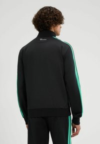 Chaqueta deportiva negra con franjas laterales verdes, cuello alto, puños acanalados y un logo en la parte posterior. Textura suave, diseño casual.