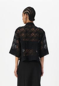 Schwarze, kurz geschnittene Jacke aus transparenter Stoff mit Rautenmuster und subtilen Logo-Akzenten. Sie hat weite Ärmel und eine gerade Silhouette.
