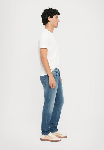 Γαλάζιο βαμβακερό φανελάκι σε εφαρμοστή γραμμή, μεσαίου μπλε τζιν παντελόνι, ελαφρά φθορά στο ύφασμα και μπεζ αθλητικά παπούτσια με πορτοκαλί λεπτομέρειες. Προφίλ από το πλάι.