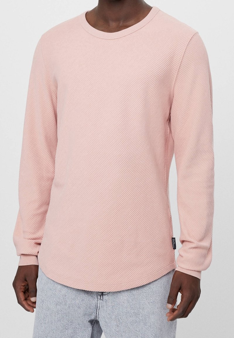 Personne portant une chemise texturée rose clair à manches longues et un jean gris clair, debout avec les bras détendus le long du corps.