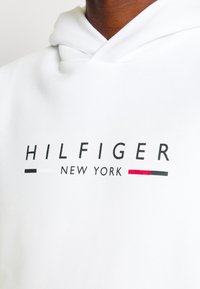 Fehér kapucnis pulóver, húzózsinórral, melyen a "HILFIGER NEW YORK" felirat áll vastag fekete és piros betűkkel. Sima anyagból készült, minimalista dizájnnal.