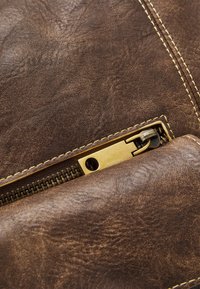 Texture en cuir marron avec grain visible, zip doré et coutures en jaune contrastant. Gros plan sur le détail du zip.