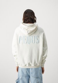 Felpa crema con tasca a marsupio frontale, caratterizzata dalla scritta "PEQUS" in azzurro chiaro sul retro. Maniche e orlo a coste.