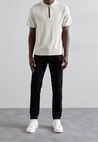 Krátký rukáv krémové polo tričko se zipovým límcem a barevným pruhem, spárované s černými džínami a bílými teniskami.