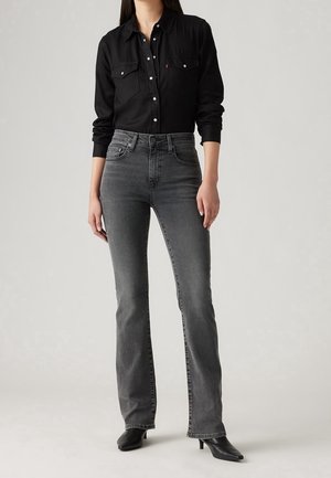 Kobieta ubrana w czarną koszulę zapinaną na guziki, szare spodnie bootcut z wysokim stanem i czarne buty na obcasie z ostrym noskiem, stojąca na szarym tle.