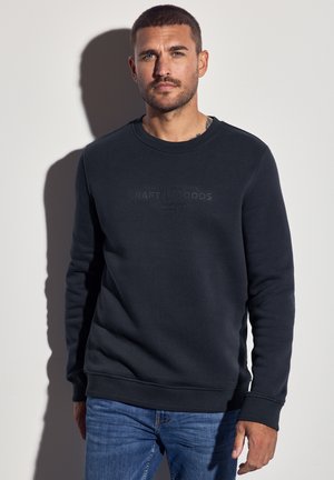 Hombre con cabello corto y barba, vistiendo una sudadera oscura con el texto "PRODUCTOS ARTESANALES" y jeans azules, de pie contra una pared blanca.