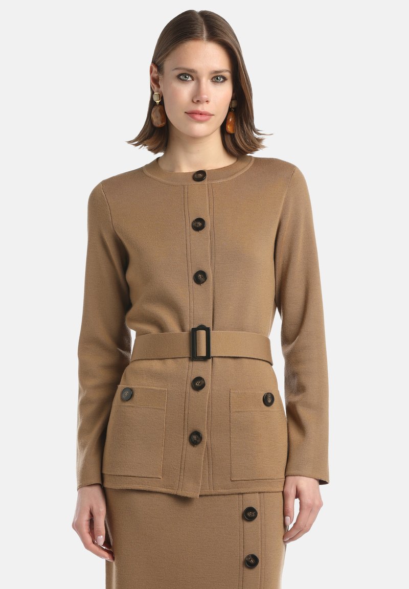 Femme portant un cardigan boutonné couleur camel avec une ceinture et une jupe assortie, accessoirisée avec de grandes boucles d'oreilles en ambre.